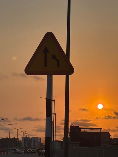 The scene captures an urban roadway in Alaguntan, Nigeria, at sunset. A prominent yellow triangular warning sign with a red border is centered in the foreground, displaying a black upward arrow and a black upward-left curving arrow, signifying a lane merge. This sign is affixed to a vertical pole marked with alternating white and red stripes. The sky exhibits a strong orange-yellow coloration, with the spherical, bright white sun visible in the lower right quadrant, partially veiled by scattered, low-lying clouds. Multiple dark, vertical poles, consistent with streetlights or utility structures, are visible throughout the midground. Along the visible horizon line, a multi-lane asphalt road contains several light-colored passenger vehicles and approximately four indistinct human figures moving or standing. The background comprises a collection of low-rise and mid-rise buildings, some appearing to be under construction. On the flat roof of a dark, multi-story building located in the mid-right, two small, dark human silhouettes are discernible.