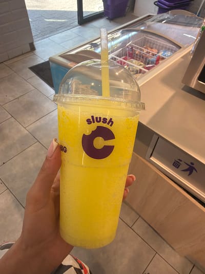 In Lviv, Ukraine, a hand with manicured fingernails holds a large, clear plastic cup filled with a yellow, slushy beverage. The cup has a domed lid and a yellow straw. Text on the cup reads "slush" in purple, with a stylized purple "C" logo. Below the logo, the word "BIG" is visible. The background shows a tiled floor, a glass-topped freezer displaying various snacks, and what appears to be a trash receptacle with a blue recycling symbol. The overall scene suggests a commercial establishment, possibly a convenience store or snack bar.