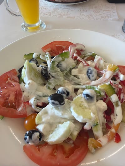 A close-up, eye-level, outdoor shot shows a white plate with a colorful salad in the foreground, positioned on a white table. The salad consists of sliced tomatoes, cucumbers, olives, and bell peppers, all coated in a creamy white dressing. A glass of orange juice is visible in the upper left corner of the image, with a red and white patterned placemat underneath the glass. The background is slightly blurred, but it appears to be a restaurant setting, possibly in the city of Sant Antoni de Portmany, Spain, given the context. No people are visible in the frame.