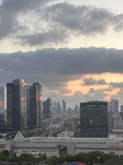 This is a cityscape view of Petah Tikva, Israel, captured from an elevated vantage point. The sky is dramatic with heavy, gray clouds interspersed with patches of lighter gray and hints of pink and orange from the setting sun.
In the foreground, several buildings are visible. Two prominent, dark glass skyscrapers, identified by signage as "B.S.R CITY," dominate the left side of the image. These buildings have horizontal bands of light running across their facades. To the right of these, another dark glass skyscraper features the "HOLMES PLACE" signage at its top, with some illuminated letters. Behind these, a denser urban landscape unfolds with numerous other buildings of varying heights and architectural styles, including some taller, slender skyscrapers in the distance.
Below the skyscrapers, a complex of lower-rise buildings forms a commercial and retail area. This includes a structure with a pyramid-shaped roof element and another large building with visible signage for various businesses, such as "ZIP," "Biga," "iDigital," and "DECATHLON." A shopping mall or complex appears to be situated here, with some signage visible, including what appears to be Hebrew text. Trees line the street in front of these commercial buildings, and cars are visible on roadways throughout the scene, indicating active city life. The overall impression is one of a modern, developed urban environment under a moody sky.