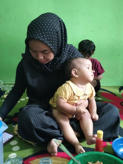An indoor scene captures a casual family moment. A woman, wearing a black hijab with white polka dots and a black long-sleeved top with thin vertical stripes, sits on a patterned floor mat with her legs crossed. She is looking downwards with a gentle expression. On her lap, she holds a baby, dressed in a yellow collared onesie, who is looking intently towards the right side of the frame. The baby has light skin and small earrings. In the background, another child with dark hair and a red shirt is partially visible, also seated on the floor.
The wall behind them is painted a bright lime green. The floor is covered with a green mat featuring colorful, abstract floral patterns in red, white, and black. Various objects are scattered around: a clear plastic jug and a smartphone with an illuminated screen are visible on the left. To the right, near the baby's feet, there is a small yellow squeeze bottle with a red cap, a blue plastic bowl, and a green plastic bowl containing food that appears to be small pieces of fried chicken or similar items, with a green utensil resting inside. A clear drinking glass is also seen in the foreground. The lighting suggests it is daytime. The overall mood is one of domestic tranquility and everyday life. No visible text is legible in the image.