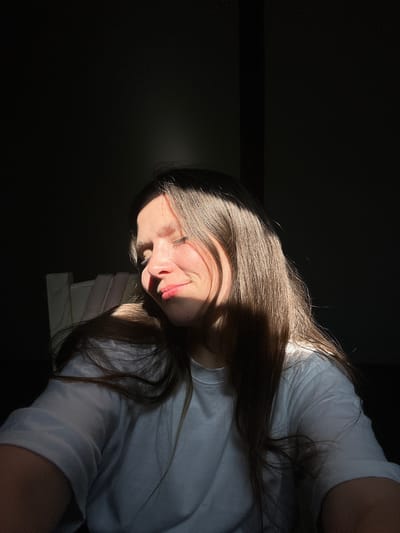 The image shows a young person, likely female, lying down in a low-light indoor setting. The photograph is rotated 90 degrees clockwise, with the person's head positioned on the right side and their body extending towards the left. A strong, directional beam of natural light, presumably sunlight, dramatically illuminates the right side of their face and their long, brown hair, creating stark contrasts with the deep shadows that envelop much of the frame and the left side of their face.
The subject is wearing a white t-shirt. Their expression features a subtle smile or smirk, suggesting contentment or relaxation. Their eyes are open, and their gaze is directed slightly upwards. Their long, flowing brown hair catches the sunlight, highlighting its texture and color. Below their head, a stack of light-colored objects, possibly books or papers, is faintly discernible, serving as a support or part of the surface they are resting on.
The background is predominantly dark, emphasizing the focused illumination on the subject. The quality of light indicates either an early morning or late afternoon setting. There is no visible text present in the image. The overall mood is intimate and tranquil, characterized by the interplay of light and shadow. While the location context is Bartula, Montenegro, no specific geographical features are visible within the image itself.