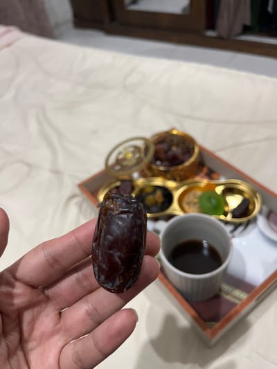 The image depicts a close-up indoor scene in Nuseirat, Palestinian Territory, focusing on a human hand holding a date. A hand with medium skin tone holds a single, dark-brown, wrinkled date fruit upright in the foreground. In the background, on a light-colored surface, possibly a bedsheet or tablecloth, a rectangular tray with a brown frame is visible. Inside the tray, an ornate gold-colored multi-compartment dish holds various items. One section contains multiple dark-brown dates, while other compartments display different dried fruits or sweets, including a prominent green-colored item. A decorative gold-colored lid with an openwork pattern rests beside the date container. To the right, a small, white or light-gray fluted cup contains a dark brown liquid, consistent with coffee. The background beyond the tray is blurred, showing a dark, wooden furniture piece.