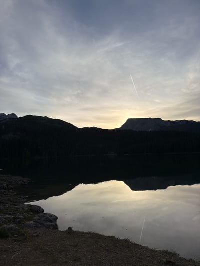 The image depicts a mountain reflected in a body of water, likely a lake, in the city of Pitomine, Montenegro. The mountain is mostly in silhouette, with a bright sky behind it, giving the appearance of a dark, inverted shape mirrored in the water. A contrail is visible in the sky, indicating an airplane. The right side of the image shows a rocky shoreline.