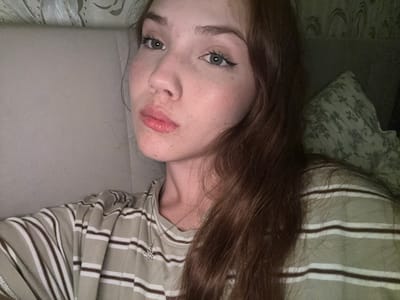 A young woman with fair skin, freckles, and auburn hair is captured in a close-up selfie. She is wearing a horizontally striped t-shirt in shades of beige and brown, with a silver pendant necklace. Her eyes are green, and she has on eyeliner. The background appears to be an indoor setting, possibly a bed or sofa, with textured wallpaper or fabric visible. The image is lit with artificial light, creating a warm glow. The location is not specified within the image itself.