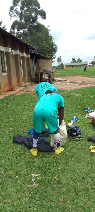 A person wearing a teal blue shirt and black shorts, number 18 on the back, and neon yellow shoes, is bending over, picking something up out of a large white bag. There is a black and white jacket lying on the green grass in front of them. Other people are visible in the background, including a person in a similar shirt but with a different number. There are several pairs of shoes scattered on the grass. There is a brown building in the background, which appears to be a residence. The location, as determined by the coordinates, is not in Lubao, Kenya, but is located in a field near the western edge of the city of Nairobi, Kenya. There is a large tree to the left of the building, and a smaller building behind the tree, which appears to be a school or sports facility.  There are many people in the background, but the majority of the image is focused on the individual in the foreground. The scene is likely taking place outdoors, in the early afternoon, as it is bright and sunny.