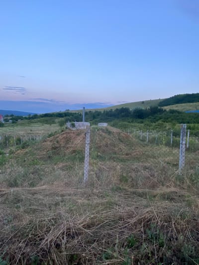 The image depicts an outdoor scene at dusk, characterized by a gradient sky transitioning from light blue to a soft twilight hue with hints of pink and lavender. In the foreground, dry, overgrown grass and hay are visible, interspersed with green vegetation. A chain-link fence with concrete posts runs across the image, indicating a boundary.
Behind the fence, a raised mound of earth, possibly a construction site or a pile of excavated material, is prominent. On top of this mound, there are concrete structures, including a circular one with a metallic pipe extending upwards, possibly a ventilation shaft or chimney. Another smaller rectangular concrete structure is also present.
In the middle ground, the landscape consists of rolling hills covered in dense green vegetation and trees. The terrain slopes upwards towards the right of the frame. A few scattered, thin poles, likely part of a fence or vineyard trellises, are visible in the grassy area.
In the background, a distant hillside with trees borders the horizon. To the far left, a red-roofed building is partially visible, suggesting a rural or semi-rural setting. A white vehicle can be seen in the distance near the building. The overall environment appears to be a developing or undeveloped area, possibly on the outskirts of a town or village, consistent with locations in and around Horodca, Moldova. The lighting suggests the transition from day to night. There are no people or animals clearly discernible in the image.