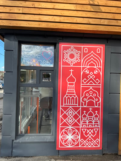 The media file shows the exterior of a building with wooden siding on the left and dark gray paneling on the right. A large red sign with white geometric patterns is mounted on the dark gray paneling. Below the sign is a window with reflections of the sky and clouds. Inside the window, neon lights spell out "БУЛКИ КОФЕ ЛЮБОВЬ" which translates to "Buns Coffee Love". A red neon heart is also visible above the text. The scene appears to be outdoors during the daytime, with natural light illuminating the wooden siding. The overall impression is that of a cafe or food establishment. The location is Vologda, Russia.