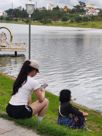 In the city of Xuân Hương - Đà Lạt, Vietnam, two individuals are observed near the lake. In the foreground, a woman wearing a white t-shirt, black shorts with white stripes, white socks, and a beige baseball cap is squatting on the grass, holding a smartphone and looking down at it. She has her hair pulled back in a ponytail and appears to be wearing earbuds. To her right, another individual, with curly dark hair, is seated on the grass facing the lake. They are wearing a dark jacket over a blue skirt and a patterned scarf draped over their shoulder with a small bag. The background features the rippling surface of Xuân Hương Lake, with a lamppost and decorative elements to the left. Further in the distance, across the lake, a tree-lined hillside is visible with buildings and structures dotting the landscape under an overcast sky.