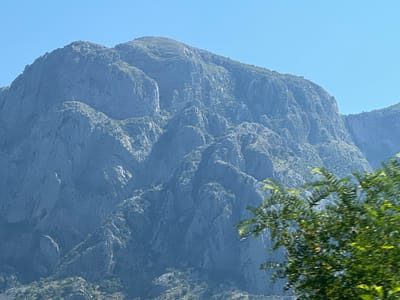 The image depicts a vast, imposing mountain range under a clear, bright blue sky, suggesting a sunny day. The mountains are rugged and rocky, with steep inclines and deep crevices. Patches of green vegetation cling to the slopes in places, hinting at a somewhat arid climate. In the foreground, a tree with vibrant green leaves is blurred due to motion, indicating the photograph was likely taken from a moving vehicle. The sunlight casts shadows on the mountains, highlighting their dramatic contours and textures. The overall scene evokes a sense of awe and the grandeur of nature. There are no visible people, objects, or text in the image. The setting is mountainous, and given the context of Škaljari, Montenegro, it is likely a view of the Lovćen mountain massif or a similar prominent peak in the region. The clear sky and bright sunlight suggest it is daytime, likely late morning or early afternoon.