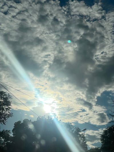 The image captures a dynamic sky filled with dramatic clouds, with the sun partially obscured by them. A bright, bluish-green circular lens flare is visible in the upper left quadrant of the image. The sun itself appears as an intense white light, creating a strong sunbeam that cuts diagonally across the frame from the upper right to the lower left, also producing a brilliant lens flare. Silhouetted against the bright sky are the dark shapes of tree branches and foliage in the upper right and lower portions of the image. Power lines stretch across the scene, further indicating an outdoor, possibly urban or suburban, setting. The sky's appearance suggests a partly cloudy day, with the sunlight breaking through in some areas, casting a warm, bright glow. The overall mood is one of natural beauty and the power of sunlight. The time of day appears to be late afternoon or early evening, given the angle of the sun and the quality of light. No people or text are visible in the image. The location context is Donskoye, Russia.