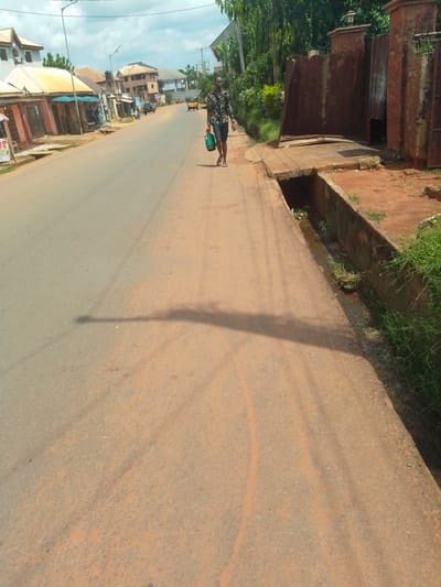 The image presents a street scene in what appears to be a town or city, consistent with Awka, Nigeria. A man walks from the background towards the foreground on the right side of the road. He has dark skin, is wearing a patterned long-sleeved shirt, dark shorts, and flip-flops, and carries a green plastic bucket in his left hand.

The road surface is predominantly a reddish-brown, possibly paved but covered in laterite dust or dirt, with visible tire tracks. A long, distinct shadow, likely from a utility pole, stretches across the road from left to right, indicating late morning or early afternoon under a partly cloudy sky. The man also casts a shadow.

On the left side of the road, several single-story buildings house shops or businesses, featuring various awnings (yellow and blue). A sign on one of these shops is partially visible, displaying "OS". Further down the street, multi-story residential or commercial buildings are seen, and a yellow tricycle (keke napep) is visible in the distance.

On the right side, a concrete drainage ditch or gutter, containing some stagnant water and vegetation, runs parallel to the road. Beyond this, a large, weathered metal gate, possibly corrugated, is set into a reddish-brown brick wall, suggesting the entrance to a private compound. Dense green foliage is visible behind this structure. Several streetlights are positioned along the road. The overall scene depicts daily life and movement in a local urban environment. The man's expression is neutral, focused on his walk.