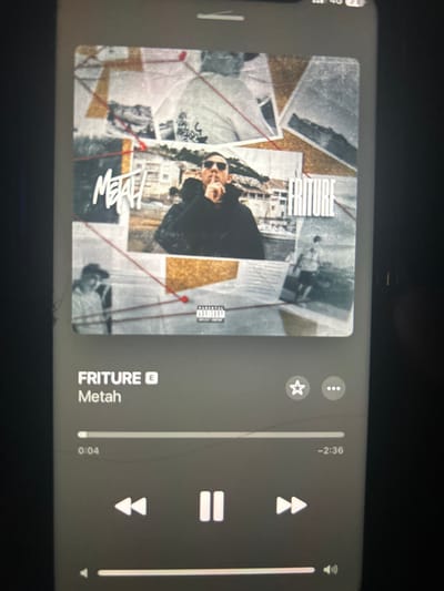 A white-framed smartphone screen, exhibiting multiple linear cracks, displays a music player interface. The screen shows the album cover for "FRITURE" by "Metah." The album cover is a collage of distressed photographs, with a central image of a man wearing sunglasses and a dark jacket, holding a finger to his lips in a "shush" gesture. This central figure is positioned against a background featuring buildings near water, possibly a coastal area. Text overlays include "MEAH" on the left and "FRITURE" on the right of the central image. A "Parental Advisory Explicit Content" label is visible at the bottom center of the cover. Other fragmented images within the collage depict individuals and landscapes. Below the album cover, the music player controls are visible, indicating the track "FRITURE" by "Metah." The playback progress bar shows 0:04 elapsed and -2:36 remaining. Pause, skip, and volume controls are present. The top right of the phone screen indicates "4G" connectivity and a battery icon. The precise physical location of this device in Bischheim, France, cannot be determined from the visual content of the image.