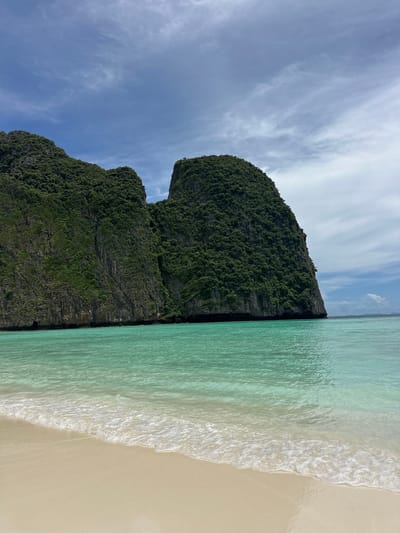 This media file captures a serene tropical beach scene in Ao Nang, Thailand. The image is oriented vertically, showcasing a dramatic limestone karst cliff covered in lush green vegetation. The cliff face dominates the left side of the frame, its rugged texture contrasted with the vibrant foliage. To the right, the clear, turquoise water of the Andaman Sea gently laps against a pristine, sandy shore. Wispy white clouds drift across a bright blue sky, suggesting a pleasant, sunny day, likely mid-morning or early afternoon given the lighting. The overall mood is tranquil and idyllic, conveying a sense of natural beauty and peacefulness. There are no people or objects visible in the image, focusing solely on the natural landscape. No text is present.