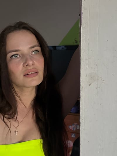 A young woman with long, dark hair and green eyes is pictured in a dimly lit room. She is wearing a neon yellow strapless top and a delicate gold cross necklace. Her mouth is slightly open, and her gaze is directed off to the left, suggesting she is looking at something or someone outside the frame. The background is blurred, but a hint of a colorful dresser with toy decals can be seen behind her. The image appears to have been taken in Rusalevka, Russia. The scene is domestic and intimate, with the woman's expression conveying a sense of curiosity or surprise.