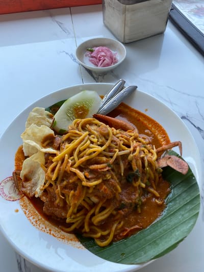 A close-up, eye-level shot shows a white plate of noodles with a red sauce, topped with bean sprouts and a whole crab. To the left of the plate are two crackers and a slice of cucumber. The plate rests on a white marble tabletop. In the background, to the right of the plate, is a small bowl of pickled red onions and a tissue dispenser. The scene is set in Lancok, Indonesia, likely at a restaurant given the presence of cutlery and a menu holder. The lighting suggests it's daytime, possibly indoors.