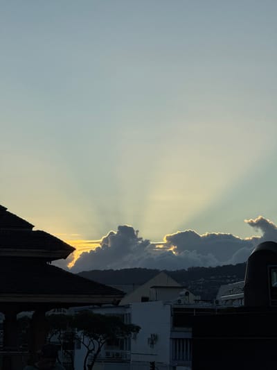 The image depicts an elevated urban view of Papeete, France, at either sunrise or sunset. The sky transitions from a light blue at the top to a pale yellow-orange gradient at the lower horizon, where the sun is obscured by dense, dark cumulus clouds. The upper edges of these clouds are brightly backlit, creating distinct crepuscular rays that ascend into the sky. A dark, forested mountain range forms the distant horizon, with a body of water possibly visible between the urban structures and the mountains.

In the lower-left quadrant, a section of a multi-tiered, dark-roofed pavilion or traditional building is prominently featured, characterized by its intricate roofline and shadowed supports. Directly below this structure, a single individual is partially visible in deep shadow, wearing a cap and positioned with their head looking downwards. The midground contains various urban buildings, exhibiting both white and dark facades, with visible windows and air conditioning units. A small tree with dark foliage is situated amongst these buildings. On the right side of the image, a dark, curvilinear architectural element, possibly a rooftop feature or part of a building, is partially visible. No explicit actions or interactions are depicted beyond the individual's posture.