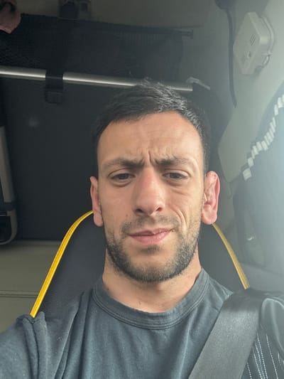The scene is located in Acconia, Italy, within the interior of what appears to be a vehicle cabin. A male subject, appearing to be in his 20s or 30s, is depicted in a close-up, chest-up shot, looking directly at the camera. He has short, dark hair and stubble covering his chin and jawline. His brow is furrowed, and his expression suggests contemplation or mild concern. He wears a dark grey or black crew-neck t-shirt. A black seatbelt is visible across his right shoulder and chest. He is seated in a black upholstered seat with a distinct vertical yellow stripe or piping along its left side. In the background to the subject's left, a dark, possibly mesh, storage pocket is visible above a horizontal silver bar. To the subject's right, a white object resembling a smoke detector or sensor is mounted on a light-colored surface, below which a dark fabric with light-colored patterned stripes is visible.