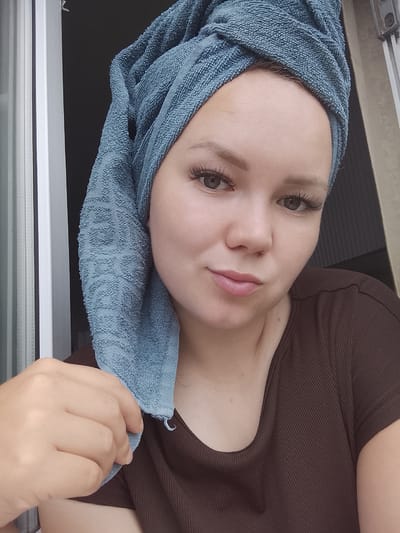A young woman with a blue towel wrapped around her head is looking at the camera. She has large, false eyelashes and her eyebrows are filled in. Her lips are slightly parted. She is wearing a brown, ribbed t-shirt. Her left hand is holding onto the towel near her shoulder. The background shows a window with blinds, suggesting an indoor setting. The lighting appears natural, possibly indicating daytime. The woman's expression is neutral, with a slight pout. There is no visible text in the image. The location is identified as Kuyeda, Russia.
