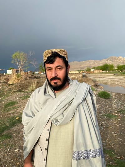 An adult male with dark hair, a full beard, and a mustache is positioned centrally, facing the camera directly. He wears a light green tunic, a buttoned beige vest, a light grey shawl draped over his shoulders with a dark blue and white geometric patterned border at one end, and an embroidered gold-colored cap. The scene is set outdoors in Arghandab, Afghanistan. The immediate ground consists of rocky, earthy terrain with sparse green vegetation. In the mid-ground, a shallow stream or canal with a muddy bed flows horizontally. Beyond the stream, the landscape includes small, partially constructed or simple buildings, scattered green trees, dry scrub, and several parked vehicles, specifically multiple white SUVs and a dark motorcycle. The distant background is dominated by a range of rugged, light-colored mountains under a sky with dark storm clouds on the left, transitioning to lighter grey towards the right.