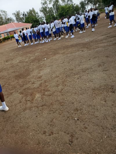 In Kura, Nigeria, a group of young males, all wearing white t-shirts and blue shorts, stand in formation on a dirt field. The individuals are positioned in several rows, facing towards the left side of the frame. Some are holding items, possibly water bottles or bags. In the background, a red-roofed building with white window frames is visible, suggesting it could be a school or an administrative facility. The surrounding environment is characterized by trees and an overcast sky. The overall scene depicts an organized gathering of students or participants.
