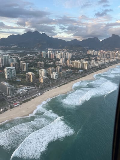 This aerial image captures a coastal urban landscape in Rio de Janeiro, Brazil, viewed from an elevated perspective, likely a helicopter or high-rise building, with a dark vertical frame element visible on the right.
The sky is overcast with a mixture of grey and lighter white-blue clouds, indicating diffuse daylight. In the distant background, a range of dark, tree-covered mountains with multiple distinct peaks spans the horizon. Some low-lying clouds are partially obscuring the upper slopes of these mountains.
In the mid-ground, an extensive urban area unfolds, featuring numerous multi-story residential and commercial buildings, predominantly light in color. These structures vary in height and architectural design. Interspersed within the cityscape are patches of green vegetation, possibly parks or undeveloped lots. A body of dark water, likely a lagoon or bay, is visible between some inland buildings and the mountain range.
The foreground is dominated by a wide, light beige sandy beach running parallel to the coastline. A multi-lane asphalt road, populated with cars and buses, separates the beach from the adjacent urban developments. Several small, red-roofed structures, consistent with beach kiosks or facilities, are visible on the sand near the road. Minimal human presence is discernible on the beach or along the road.
The Atlantic Ocean occupies the lower and right portions of the image, displaying shades of turquoise and deep blue. Multiple parallel lines of white-crested waves are actively breaking and rolling towards the shore, forming distinct patterns of foam and whitewater. The overall scene illustrates a developed coastal zone at the base of substantial mountain ranges, characteristic of the Barra da Tijuca or Recreio dos Bandeirantes areas of Rio de Janeiro.