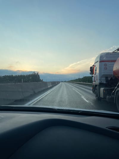The image is taken from inside a vehicle, looking out at a highway at dusk. The sky is a gradient of light blue and yellow, with wispy clouds. The road ahead is wet, reflecting the light. To the right of the road is a white semi-truck with red trim. On the side of the truck, there are circular signs indicating speed limits of 90 and 70. The truck is partially obscured by the edge of the frame. On the left side of the road, there is a concrete barrier, and beyond that, a line of trees and hills. The interior of the vehicle, specifically the dashboard and windshield, are visible in the foreground, indicating the perspective is from the driver's seat. The location is identified as Novozhilovo, Russia.