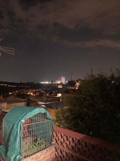 The image captures an urban nighttime scene, likely from a rooftop or elevated vantage point, looking out over a town or city. In the foreground, a structure covered with a green tarp and enclosed by a metal grate is visible. Beyond this, rooftops of various buildings spread out, some illuminated by interior lights. In the middle ground, a distant cityscape is dotted with lights, and a taller, multi-story building with illuminated windows stands out. The sky above is dark with scattered clouds, suggesting evening or nighttime. The presence of a windmill-like structure with illuminated elements in the far left background, combined with the general architecture, suggests the location could be in or near Sabadell, Spain, a city known for its industrial past and some preserved windmills.