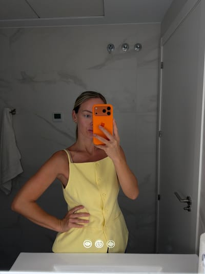 A woman is taking a mirror selfie in what appears to be a bathroom. She is wearing a yellow sleeveless top and has her right hand on her hip. She is holding an orange iPhone in her left hand, which is angled to capture her reflection. The background shows a marble-tiled wall, a towel hanging on a rack, a door with a handle, and a sink with illuminated touch controls. The overall impression is that of a modern, well-lit bathroom. There is no visible indication of the specific city or country.
