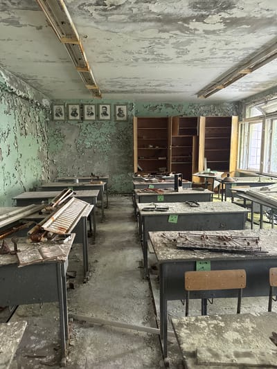 The image depicts a deteriorated room in Pryp'yat', Ukraine, characterized by peeling paint on the walls and floor. The left wall has a row of framed portraits and a decaying electrical fixture. A wooden shelving unit is against the back wall. The right side features a series of metal boxes with attached apparatuses along the wall. The room is illuminated by natural light entering through a window in the ceiling.