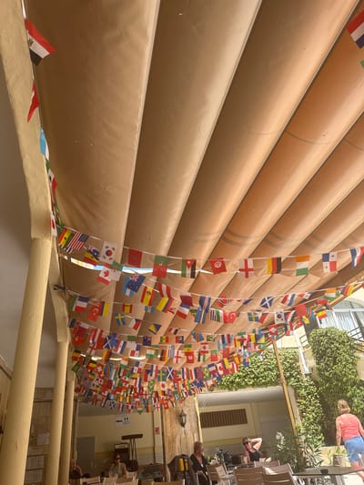 The image depicts an outdoor seating area with numerous flags of various countries strung across the ceiling under a tan awning. The flags are arranged in multiple rows, creating a festive atmosphere. In the background, there is a building covered in lush green ivy, and several people are seated at tables, appearing to be patrons of a cafe or restaurant. One woman in a pink top and blue shorts is standing, facing away from the camera. The scene is situated in the city of Fuengirola, Spain, suggested by the architecture and the warm, sunny weather. The flags likely represent the diversity of visitors or a connection to global events.