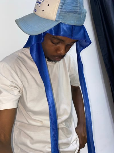 A man is seen sleeping. He is wearing a light blue hat and a blue satin wrap. He is lying on his back, wearing a white t-shirt. His eyes are closed. His hand is on the right side of his body. The background is white and black, with a shadow in the top corner.