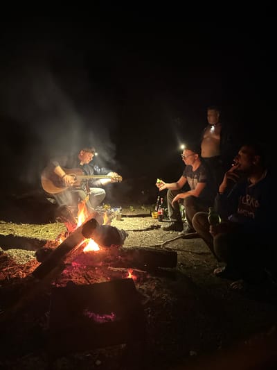The image shows a nighttime scene with a group of people gathered around a campfire. One man is playing an acoustic guitar. Two other men are wearing headlamps. The location is Юзуфоўскі сельскі Савет, Belarus.
