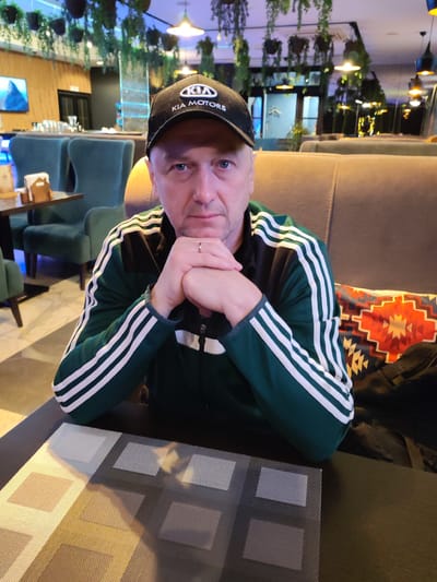 A middle-aged Caucasian male is seated at a table in what appears to be a restaurant or cafe. He is wearing a dark green jacket with white stripes on the sleeves and a black baseball cap with a "KIA MOTORS" logo. His hands are clasped in front of his face, resting on the table. The background suggests an indoor setting with a dimly lit ambiance, possibly enhanced by decorative lighting and hanging plants. A table setting is visible in front of him, featuring a textured placemat. The environment appears to be a public space with other seating areas and decor visible. There are no immediate indications of the specific location within Yoshkar-Ola, Russia.