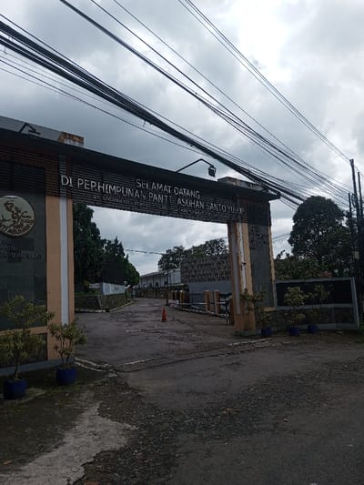 The image displays the entrance to an institutional facility, captured under an overcast sky. A prominent archway, constructed with light orange-brown and dark grey stone or tile, spans an asphalt road. The archway's main signage reads "SELAMAT DATANG DI PERHIMPUNAN PANTI ASUHAN SANTO YUSUP" (Welcome to the Association of Saint Joseph Orphanage) in white letters, with "JL. SINDANGJAYA CIJANTUNG" (Sindangjaya Cijantung Street) visible below, indicating a location in Indonesia. The left pillar features a circular emblem and additional text, "MELAYANI ANAK BANGSA, MENCINTAI ANAK ALLAH." The right pillar contains obscured text including "PEMERINTAH" and "DINAS SOSIAL."
The asphalt road, showing signs of wear and repair, leads inward past the archway, curving slightly to the right towards distant buildings. An orange traffic cone is positioned on the road within the entrance area. Various trees and shrubs are visible around the entrance, with small potted plants placed near the base of the archway pillars. Overhead, numerous black utility lines traverse the upper frame, connecting to a pole on the right. The sky is uniformly grey and cloudy. No individuals or vehicles are present. The explicit Indonesian street address on the sign contradicts the specified location of Gubbisamamidi, India.