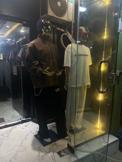 This image depicts a retail storefront located in Iranla, Nigeria, displaying two mannequins through a glass partition. The mannequin on the left has a gold-toned head and hands, wearing a dark blue beanie embellished with multi-colored circular studs, a brown zippered jacket, and black pants with a gold crown logo near the left hip, from which a silver chain dangles. This mannequin stands on a blue mat, wearing white footwear. The mannequin on the right, also with a gold-toned head and hands, wears a red and white baseball cap, an off-white short-sleeved t-shirt bearing the word "LIAISON" in dark grey letters, and matching off-white pants. This mannequin stands on a dark mat, wearing dark footwear.
Behind the mannequins, visible through another glass partition, is the store interior with racks of clothing and bright overhead lighting. Above the mannequins, a white Hisense brand air conditioning unit is mounted, with exposed electrical conduits and wiring. The exterior floor is covered with white marble-patterned tiles. To the right of the glass door, a green wall is visible, featuring a vertical yellow LED strip light, a chrome door handle, and a small white sign with partial text "0814..." affixed to it.