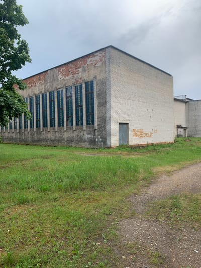 An outdoor, eye-level, wide-angle shot captures the side of a light brick building with a large expanse of glass block windows running along its length. The building has a weathered appearance, with exposed red brick and concrete sections visible at the top and on the left side. A small, grey door is set into the brick wall on the right side of the building. The building is situated in a grassy area with a gravel path leading away from it. A tree with green leaves is visible in the upper left corner of the frame. The sky is overcast with clouds. The scene is devoid of people or any observable activity. The architectural style suggests it may be an industrial or institutional building.