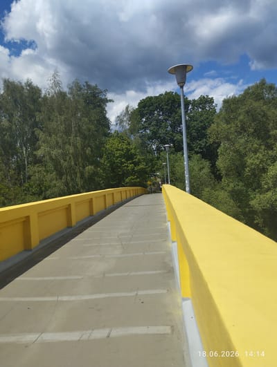 The image captures a pedestrian bridge with bright yellow railings, curving to the right as it ascends. The bridge's walkway is paved with concrete and marked with faint white lines. Two streetlights are visible along the bridge, with one closer to the foreground and another further down the path. Dense green trees flank the bridge on both sides, forming a natural barrier. Above, a dynamic sky displays a mix of dark storm clouds and patches of bright blue sky. The lower right corner of the image features a timestamp: "18.06.2026 14:14". The setting appears to be an outdoor urban environment, likely in Karlovy Vary, Czechia, given the presence of a pedestrian bridge and surrounding greenery.
