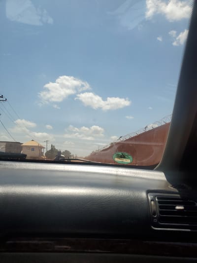 The image depicts a bright daytime scene in Yelwa, Nigeria, viewed from inside a vehicle through its windshield. The foreground features a dark-colored dashboard, reflecting sunlight, with a visible air vent on the far right. The sky is clear blue with scattered white cumulus clouds.
On the left side of the exterior view, utility poles with multiple overhead wires are present. A light-colored building with an orange-brown pitched roof and a veranda stands, with a plainer concrete structure visible behind it, along with distant trees. A dark, indistinct shape, possibly a person on a motorcycle or a stationary object, is visible in the middle ground, partially obscured by reflections on the windshield.
On the right side of the exterior view, a long, reddish-brown wall topped with razor wire extends into the distance, indicating a secure perimeter. A green oval sticker with the text "TRD82 TX" is affixed to the interior of the windshield, partially overlapping the view of the wall. The road surface in the middle distance appears unpaved and dusty. Reflections, possibly of the vehicle's interior or the sky, are visible on the upper left portion of the windshield.