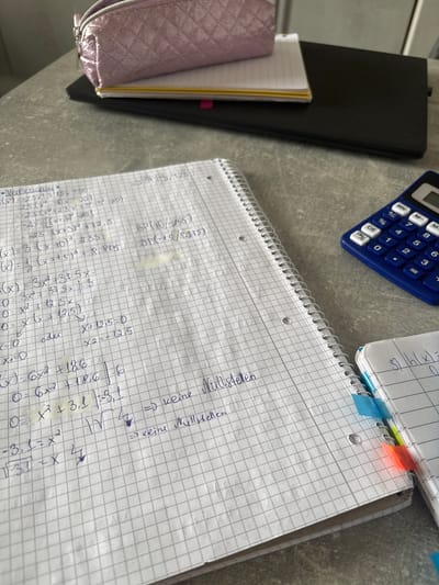 The image depicts a close-up, eye-level view of a cluttered desk surface, likely in a home or educational setting. The desk is made of a gray, textured material. Dominating the foreground is a spiral-bound notebook, open to pages filled with handwritten mathematical equations and calculations. The writing is in blue ink and appears to be in German. Several sticky notes in blue, green, and orange are attached to the edge of the notebook.

To the right of the notebook is a blue calculator with a white keypad. To the left and slightly behind the notebook, a pink, sparkly pencil case rests on top of a black object, possibly a laptop or a binder. The pencil case is partially open, revealing a stack of papers or another notebook underneath.

The background shows more of the gray desk surface and what appears to be a light-colored wall or divider. There are no people visible in the image. The lighting suggests an indoor environment, possibly with natural light from a window.

The scene does not provide any direct information about the specific location within Groß-Gerau, Germany. The objects and the content of the notebook (mathematical work) suggest an academic or study-related context.