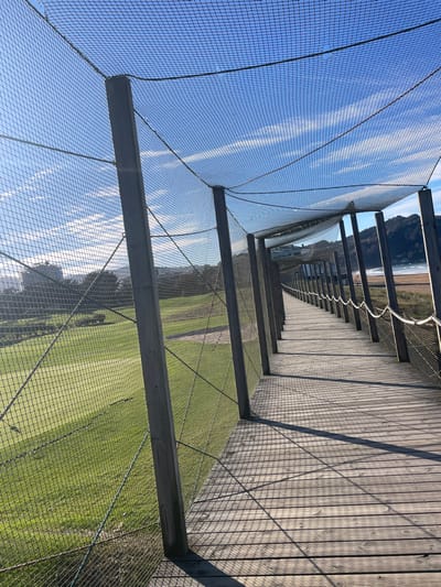 The image displays a linear wooden boardwalk, enclosed and partially covered by a tall protective netting structure supported by numerous wooden posts. The netting casts a distinct grid-like shadow pattern onto the sunlit, weathered wooden planks of the path. To the immediate left, through the netting, a meticulously maintained green golf course is visible, featuring fairways and sand traps. To the right of the boardwalk, a sandy beach extends towards the ocean, where gentle waves are observed. In the background, distant hills covered in vegetation frame the coastline, and scattered urban buildings are discernible beyond the golf course. The sky is bright blue with scattered wispy white clouds. The scene is illuminated by strong sunlight from the left, indicating a clear day. No individuals are present. This structure functions as a pedestrian walkway separating the Zarautz Golf Course from Zarautz Beach in Zarautz, Spain.