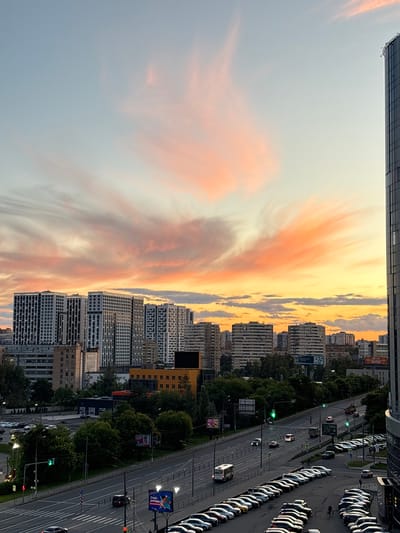 This is a landscape photograph taken from a high vantage point, likely a building, overlooking a city street and residential buildings at dusk. The sky is filled with dramatic, wispy clouds painted in hues of orange, pink, and yellow, indicating a sunset.

The scene features a multi-lane road with several cars driving and a long line of parked cars on the right side of the road. Traffic lights are visible, with some showing green. Trees line the street, interspersed with modern residential buildings, some of which are tall and have many windows. In the foreground, a portion of a modern building with a dark facade is visible at the top edge of the frame.

There are several visible signs. One sign on the right side of the road features a colorful advertisement with a woman and text. Another sign near the road is a billboard. Smaller signs and traffic symbols are also present throughout the scene.

The overall mood of the image is serene and picturesque, capturing the transition from day to night with the beautiful sunset. The location is identified as Mytishchi, Russia.