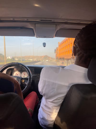 The image shows the interior of a vehicle, specifically the driver's seat. The viewpoint is from the passenger seat. A dark-skinned person with braided hair, wearing a white shirt and a black jacket, is partially visible in the driver's seat. The driver's hands are on the steering wheel. The dashboard is visible, with gauges and controls. The windshield has a crack. Through the windshield, a view of Jiru, Nigeria, is visible. An orange building is prominent in the background. The sky is light.