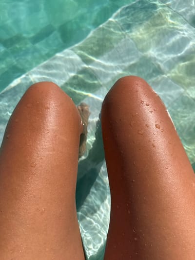 The image displays a close-up, top-down view of two human legs, exhibiting a tanned complexion, partially submerged in a swimming pool in Tivat, Montenegro. Numerous clear water droplets are visible on the sun-kissed epidermal surface of both legs. The pool water is distinctly turquoise, with patterns of refracted sunlight dancing across its surface and illuminating the submerged white or light-colored square tiles on the pool floor. A blurred outline of human feet is discernible further down in the water.