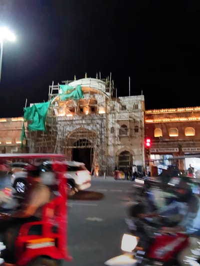 The image shows Hawa Mahal (Palace of Winds) located in Jaipur, India at night. The building is under construction, with scaffolding covering the facade and green banners hanging from the structure. There are buildings in the background, and traffic moving in front of the monument. The night sky is dark.