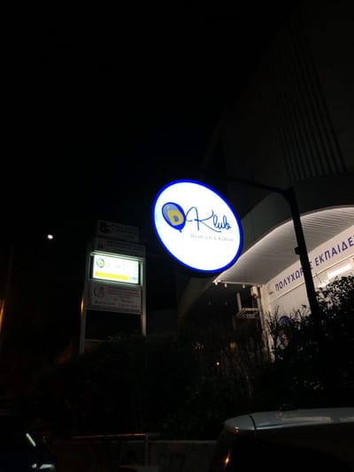 The image depicts a nighttime urban scene in Marousi, Greece, illuminated primarily by artificial lights. The central subject is a brightly glowing, oval-shaped sign suspended from a dark pole. The sign is white with a blue border and features the stylized text "O Klub" above "Deutsch & Kultur". The letter "O" in "O Klub" is replaced by a distinctive logo resembling an "evil eye" or a balloon with an eye motif, colored yellow, blue, and white.
Behind and above the main sign, a modern building facade with vertical slats is partially visible. Inverted text on the building reads "ΠΟΛΥΧΩΡΟΣ ΕΚΠΑΙΔΕΥΣΗΣ" (Polyhoros Ekpaidefsis), which translates to "Multipurpose Educational Space."
Below the main sign, a multi-panel street sign board is also illuminated, displaying several smaller signs. One of these, with a yellow background, clearly shows the same eye logo seen on the main sign, alongside Greek text that includes "ΒΡΕΦΟΝΗΠΙΑΚΟΣ ΣΤΑΘΜΟΣ" (Infant/Toddler Station/Nursery). Another panel mentions "2ος Όροφος" (2nd Floor), and another sign appears to reference "BEBE DUCK".
The remainder of the scene is dark, with silhouetted trees or bushes on the right side and a small, bright point of light visible lower down. A part of a dark-colored vehicle is just visible on the far right edge. No people are present, lending the scene a quiet, late-night atmosphere.