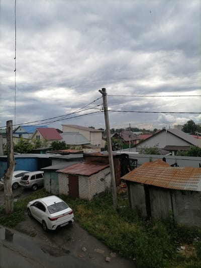 The image depicts a cloudy day over a residential area, possibly a dacha cooperative or a district of private homes and garages in Ulyanovsk, Russia. In the foreground, a white sedan is parked on a dirt and asphalt surface. To the left, two other vehicles, a white hatchback and a silver station wagon, are parked in front of blue and grey garages. A dilapidated wooden pole with electrical wires runs vertically along the left side of the frame. To the right, several garages with corrugated metal roofs are visible, one of which appears to be made of brick and has a rusty metal roof with an open door. In the background, various houses with pitched roofs of different colors, including blue and red, are situated amongst trees. The sky is heavily overcast with dense grey clouds, suggesting a possibility of rain.