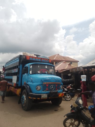 The image depicts a street scene in Serti, Nigeria, dominated by a bright blue truck adorned with colorful decorations and the text "THANK GOD" in large letters across its top. The truck is loaded with goods, some covered with tarpaulin. In the foreground, a person wearing a plaid shirt and shorts walks away from the camera. To the right, a motorcycle is parked, and a person in a purple shirt is visible. Further back, another motorcycle and a woman in a red headscarf carrying a bucket on her head are present. The background features buildings with rusty, corrugated iron roofs, indicative of the local architecture. The sky is partly cloudy, with large, white clouds dominating the upper portion of the image. The scene appears to be a typical roadside setting in a Nigerian town, possibly a market area.