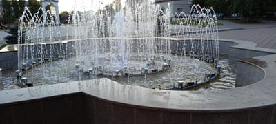 This media file shows a fountain in an outdoor setting. The fountain features multiple water jets spraying upwards, creating a curtain of water that catches the light. Lights are visible beneath the water's surface, casting colorful reflections. In the background, a building with a light-colored facade and windows can be seen, along with trees and paved walkways. The scene appears to be in a public space, possibly a park or town square, given the presence of benches and the architectural elements. The lighting suggests it is daytime, with soft sunlight filtering through the water and illuminating the scene. The overall impression is one of a pleasant, serene environment, likely during a calm weather day. There is no visible text in the media file.
