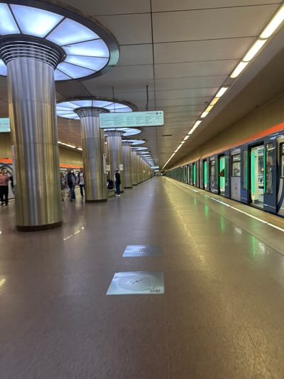 The image depicts the interior of a modern, well-lit metro station, likely in Kotelniki, Russia. The perspective is from a platform looking down a long tunnel. The station features a sleek design with a light-colored tiled wall on the left and a textured, reflective floor on the right. Large, cylindrical metal columns with illuminated circular accents line the platform. A train is visible in the distance, partially obscured by the perspective and lighting.
Several people are visible on the platform, appearing to be waiting for or disembarking from a train. Their activity suggests a typical transit scene. The lighting is bright and uniform, indicating it is likely daytime or the station is artificially illuminated, which is common for subway environments. The overall atmosphere appears calm and functional.
Notable details include the clean lines of the architecture, the futuristic design of the columns, and the reflective surfaces that amplify the station's lighting. There are signs visible on the columns, though the text is not legible at this resolution. Two square panels are mounted on the wall to the right, one with a grille pattern and another with what appears to be a clock or informational display, also not clearly readable. The presence of a metro train and the style of the station architecture are strong cues to its urban, public transportation setting.